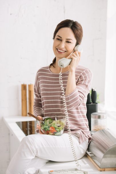 smiling woman talking on retro phone while sitting on kitchen table and holding bowl with vegetable salad - Foto, immagini