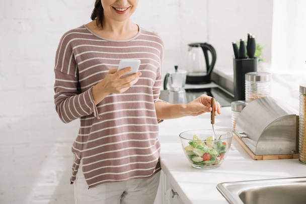 vista recortada de la mujer sonriente usando un teléfono inteligente mientras está de pie en la mesa de la cocina cerca del tazón con ensalada de verduras
 - Foto, imagen