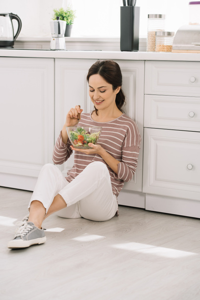 νεαρή, χαρούμενη γυναίκα που κάθεται στο πάτωμα της κουζίνας και τρώει σαλάτα λαχανικών - Φωτογραφία, εικόνα
