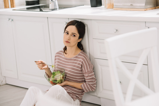 dissatisfied young woman sitting on floor in kitchen and eating vegetable salad - Photo, Image