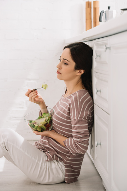 upset young woman sitting on floor in kitchen and eating vegetable salad - Foto, Imagen