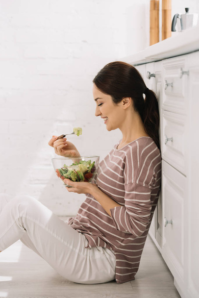 heureuse jeune femme assise sur le sol dans la cuisine et manger de la salade de légumes
 - Photo, image