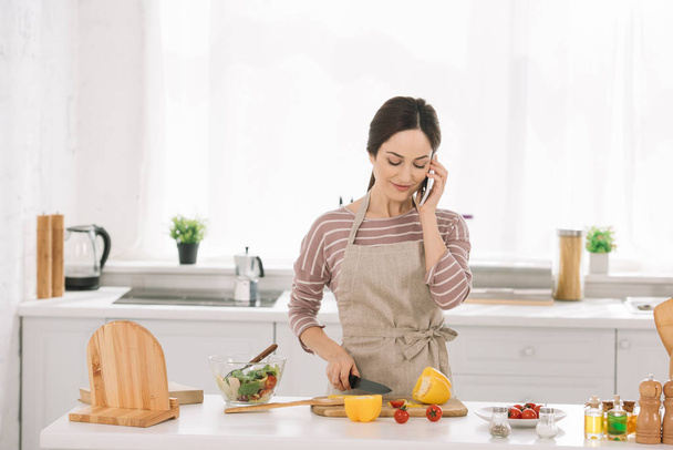 smiling woman in apron cutting bell pepper while standing near kitchen table with vegetables and talking on smartphone - Φωτογραφία, εικόνα