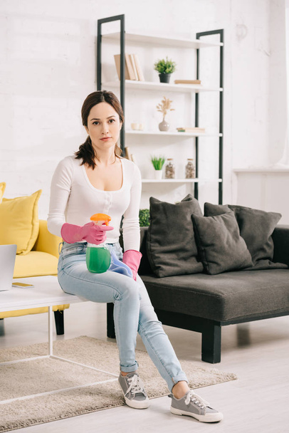 attractive housewife looking at camera while sitting on table and holding rag and spray bottle - Foto, afbeelding