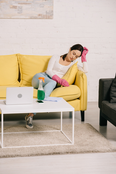 dona de casa cansada sentado no sofá amarelo com os olhos fechados perto da mesa com laptop e spray garrafa
 - Foto, Imagem