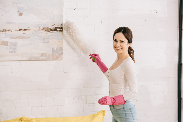 cheerful housewife smiling at camera while cleaning picture on wall with dusting brush - Photo, Image