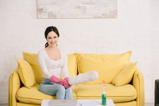 happy housewife smiling at camera while sitting on yellow sofa and holding dusting brush - Fotografie, Obrázek