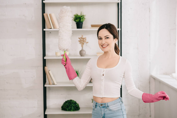 young, cheerful housewife looking at camera while holding dusting brush - Photo, Image
