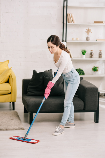 young housewife washing floor with mop near grey sofa - Photo, image