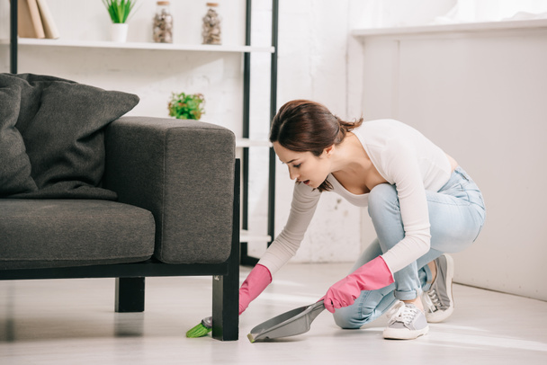 junge Hausfrau fegt Fußboden neben Sofa mit Pinsel und Schaufel - Foto, Bild