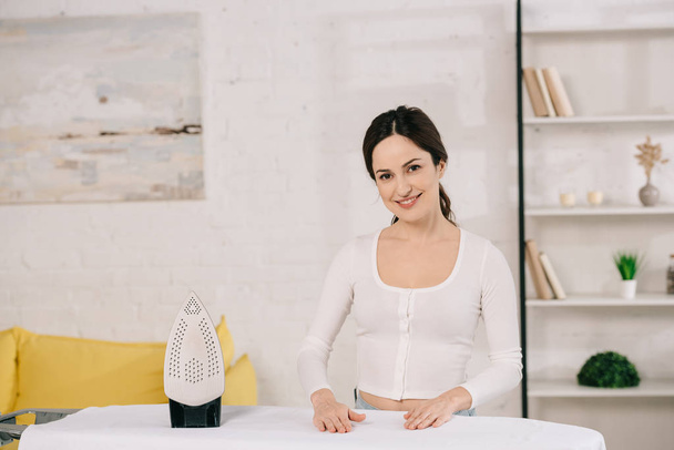 happy housewife smiling at camera while standing near ironing board - Valokuva, kuva