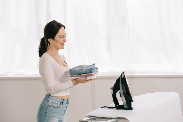 happy housewife holding ironed clothes while standing near ironing board - Foto, Bild