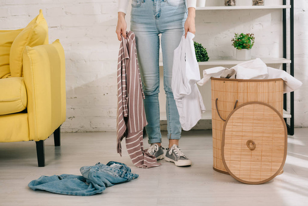 oříznutý pohled na hospodyni v džínách džínách držící oblečení při stání v blízkosti prádelního koše - Fotografie, Obrázek