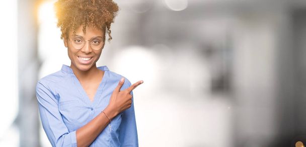 Mooie jonge Afro-Amerikaanse zakenvrouw over geïsoleerde achtergrond vrolijke met een glimlach van het gezicht wijzen met hand en de vinger aan de kant met gelukkig en natuurlijke uitdrukking op het gezicht - Foto, afbeelding