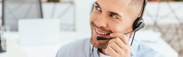 panoramic shot of happy and handsome operator in brokers agency touching headset  - Фото, изображение