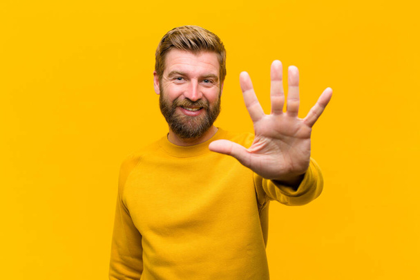 young blonde man smiling and looking friendly, showing number five or fifth with hand forward, counting down against orange wall - Foto, Imagen
