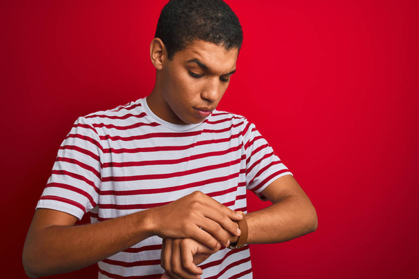 Młody przystojny arabski mężczyzna w pasiastej koszulce na izolowanym czerwonym tle Sprawdzanie czasu na zegarku, zrelaksowany i pewny siebie - Zdjęcie, obraz