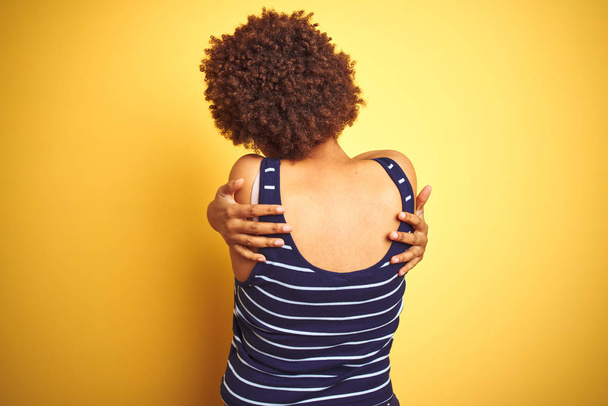 Beauitul Afrikaanse Amerikaanse vrouw draagt zomer t-shirt over geïsoleerde gele achtergrond knuffelen zich gelukkig en positief van achteren. Zelfliefde en zelfzorg - Foto, afbeelding