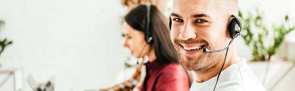πανοραμική φωτογραφία του χαρούμενου μεσίτη σε ακουστικά που εργάζονται στο γραφείο με συναδέλφους  - Φωτογραφία, εικόνα