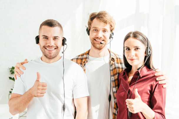 happy brokers in headsets looking at camera and showing thumbs up  - Photo, image