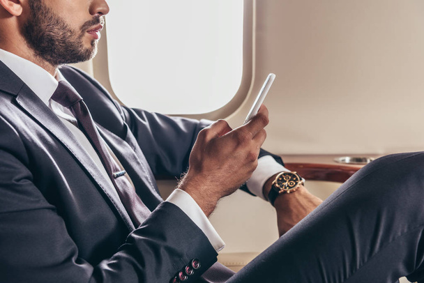 oříznutý pohled na podnikatele v obleku pomocí smartphonu v soukromém letadle  - Fotografie, Obrázek