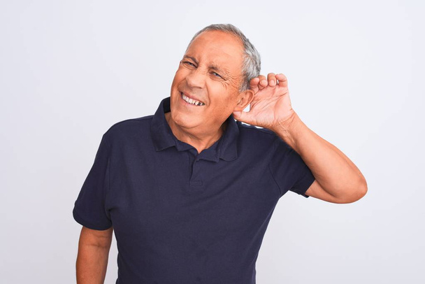 Homem de cabelos grisalhos sênior vestindo pólo casual preto em pé sobre fundo branco isolado sorrindo com a mão sobre o ouvido ouvindo uma audição a rumores ou fofocas. Conceito de surdez
. - Foto, Imagem
