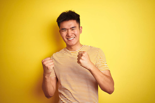 Jonge Aziatische Chinese man die een t-shirt draagt en over een geïsoleerde gele achtergrond staat, is erg blij en enthousiast en doet winnaarsgebaar met opgeheven armen, glimlachend en schreeuwend om succes. Feestconcept. - Foto, afbeelding