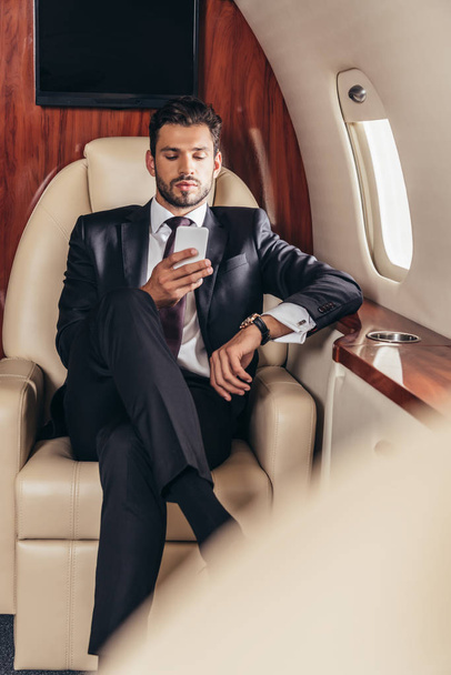 избирательный фокус привлекательного бизнесмена в костюме с использованием смартфона в частном самолете
  - Фото, изображение