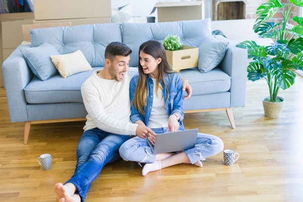 Young couple sitting on the floor of new home using computer laptop and smiling happy for moving to a new home - Photo, Image