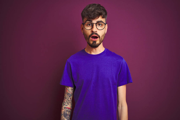 Fiatal férfi tetovált visel póló és szemüveg felett áll elszigetelt lila háttér fél, és sokkolt meglepő kifejezés, félelem és izgatott arc. - Fotó, kép