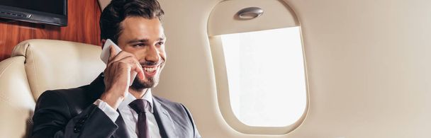 πανοραμική λήψη του όμορφου επιχειρηματία με κοστούμι χαμογελώντας και μιλώντας στο smartphone σε ιδιωτικό αεροπλάνο  - Φωτογραφία, εικόνα