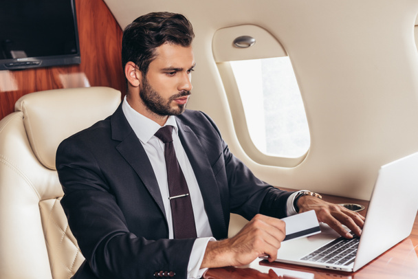 schöner Geschäftsmann im Anzug mit Kreditkarte und Laptop im Privatflugzeug  - Foto, Bild
