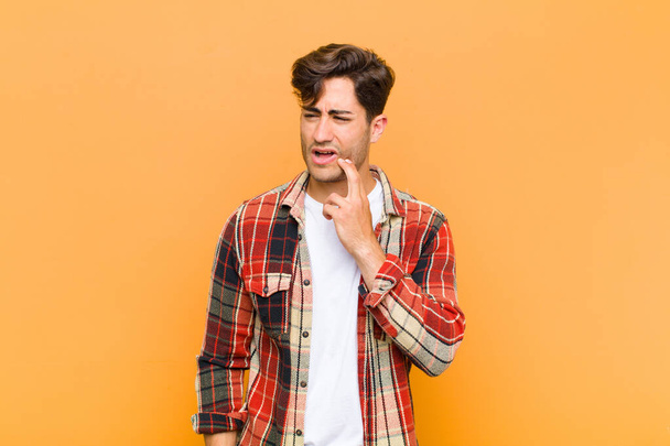 young handsome man holding cheek and suffering painful toothache, feeling ill, miserable and unhappy, looking for a dentist against orange background - 写真・画像
