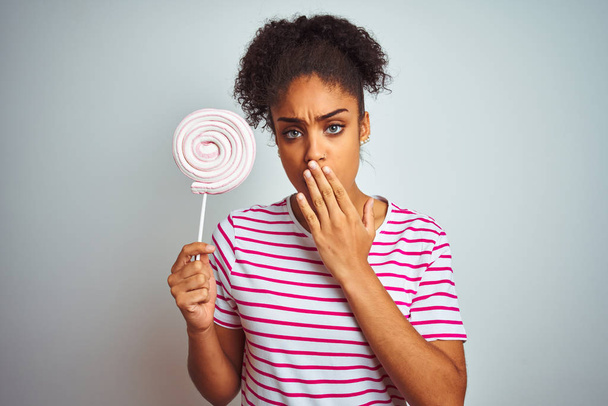 Afrykańska nastolatka jedząca kolorowe cukierki nad odizolowanym białym tłem usta z ręką zaszokowaną wstydem przez pomyłkę, wyrażeniem strachu, przestraszoną w ciszy, sekretną koncepcją - Zdjęcie, obraz