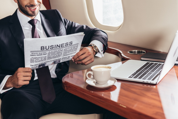 visão recortada de empresário sorridente em terno ler jornal de negócios em avião privado
  - Foto, Imagem