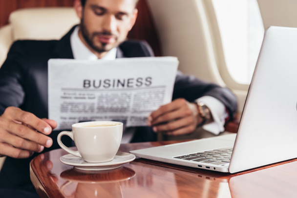 επιλεκτική εστίαση του επιχειρηματία στο κοστούμι ανάγνωση επιχειρηματική εφημερίδα και τη λήψη κύπελλο στο ιδιωτικό αεροπλάνο  - Φωτογραφία, εικόνα