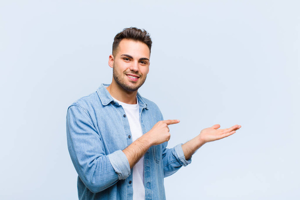 young hispanic man smiling, feeling happy, carefree and satisfied, pointing to concept or idea on copy space on the side against blue wall - Fotó, kép