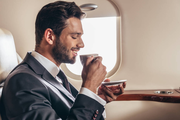 boční pohled na usmívajícího se podnikatele v obleku drží šálek kávy v soukromém letadle  - Fotografie, Obrázek