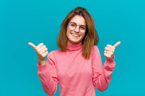 young pretty woman smiling joyfully and looking happy, feeling carefree and positive with both thumbs up against blue background - 写真・画像