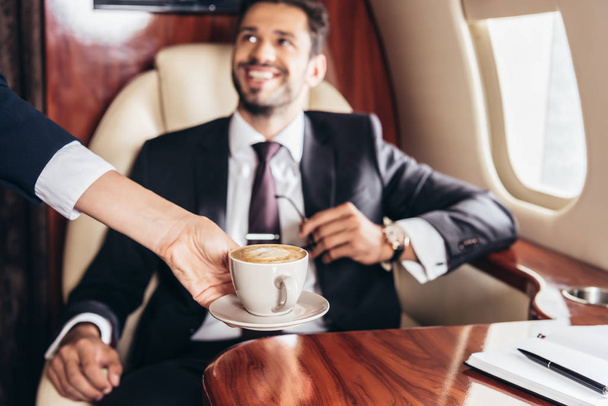 Uçuş görevlisinin özel uçaktaki takım elbiseli yakışıklı iş adamına kahve verirken çekilmiş görüntüsü.  - Fotoğraf, Görsel