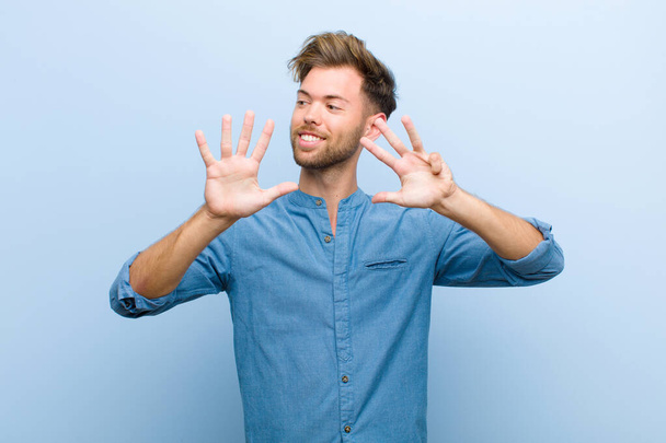 young businessman smiling and looking friendly, showing number nine or ninth with hand forward, counting down against blue background - Foto, afbeelding
