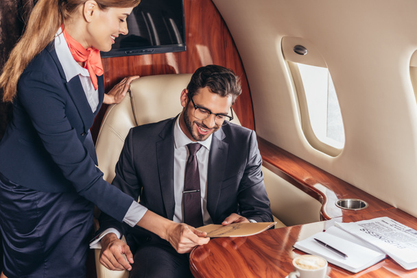 Flugbegleiterin gibt lächelndem Geschäftsmann im Anzug im Privatflugzeug Menü  - Foto, Bild