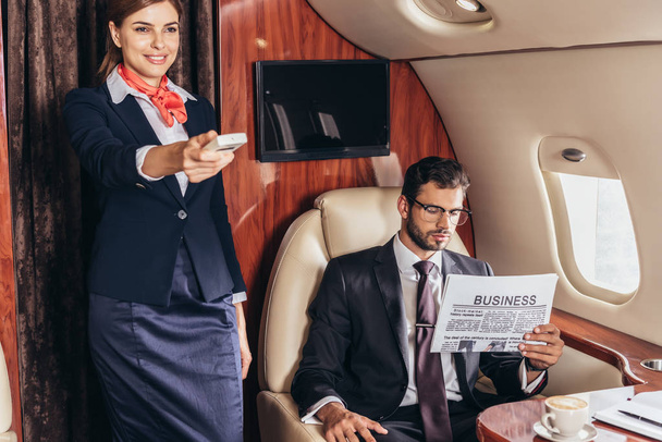 schöner Geschäftsmann im Anzug liest Zeitung und lächelnder Flugbegleiter hält Fernbedienung im Privatflugzeug  - Foto, Bild
