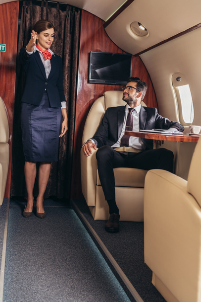 Flugbegleiterin zeigt im Privatflugzeug Geste gegenüber hübschem Geschäftsmann im Anzug  - Foto, Bild