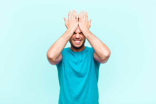 jonge Spaanse man voelt zich gestrest en angstig, depressief en gefrustreerd door hoofdpijn, het verhogen van beide handen naar het hoofd tegen een blauwe achtergrond - Foto, afbeelding