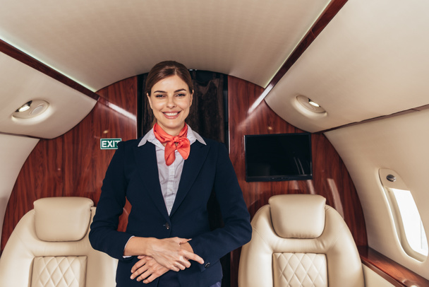 smiling flight attendant in uniform looking at camera in private plane  - Fotografie, Obrázek