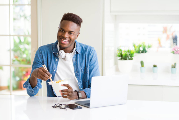 Komea nuori afrikkalainen liikemies syö toimitusta aasialainen ruoka ja työskentelee tietokoneella, nauttien nuudeleista hymyillen
 - Valokuva, kuva