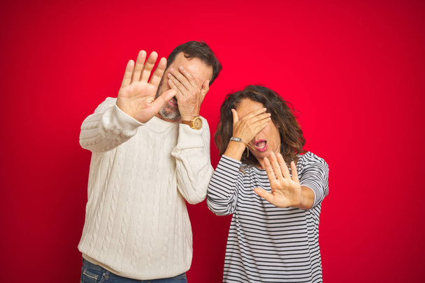 Beautiful middle age couple wearing winter sweater over isolated red background covering eyes with hands and doing stop gesture with sad and fear expression. Embarrassed and negative concept. - Фото, изображение