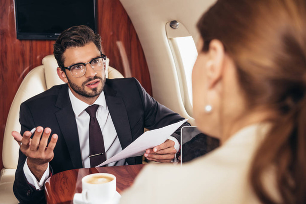 selectieve focus van zakenman die naar zakenvrouw kijkt en papier in privévliegtuig houdt  - Foto, afbeelding