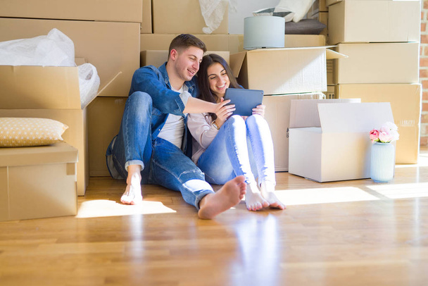 junges Paar sitzt auf dem Boden des neuen Hauses um Pappkartons mit Tablet herum und lächelt glücklich für neue Wohnung - Foto, Bild